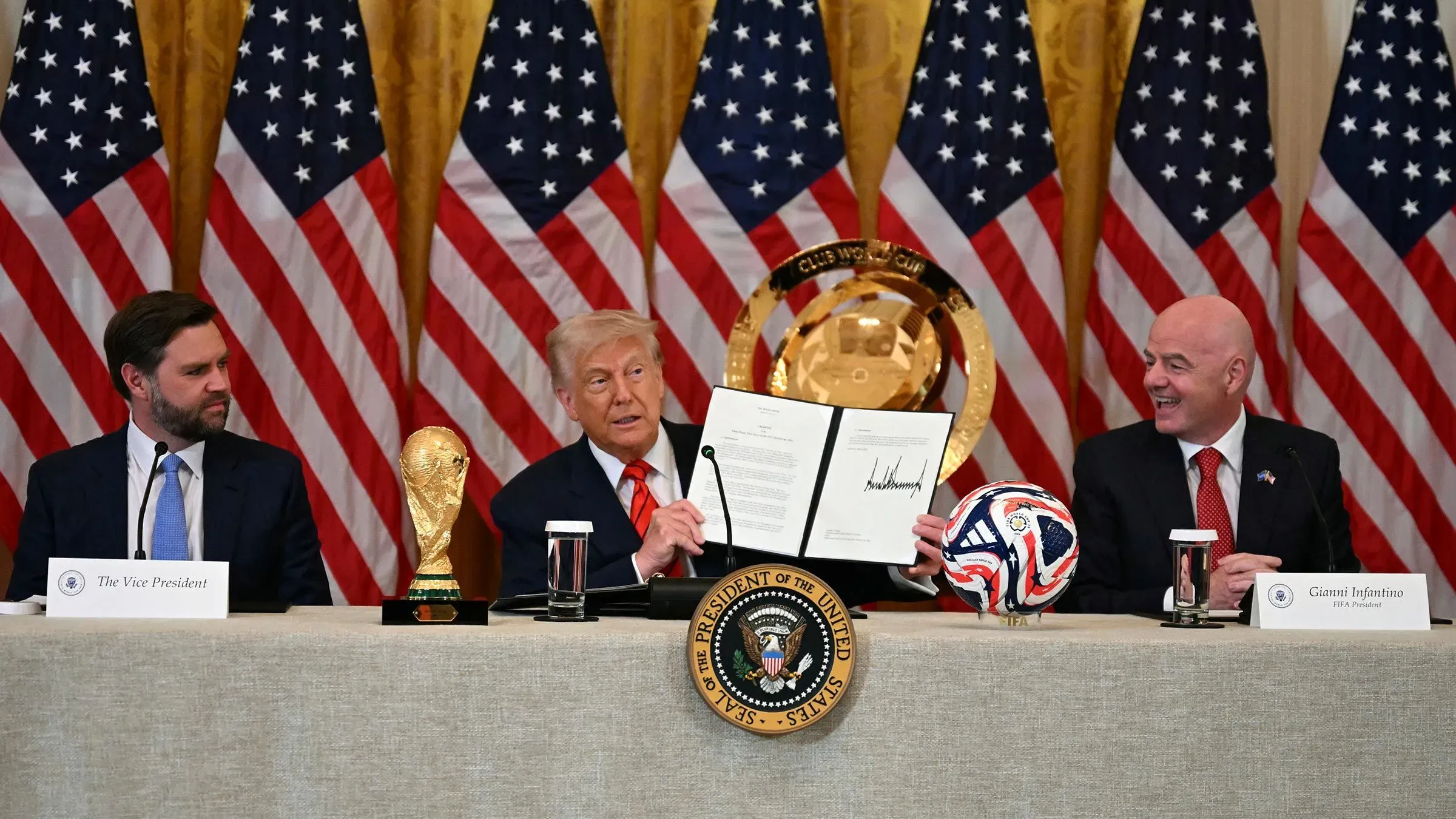 ترامب يتحدث خلال اجتماع حول كأس العالم 2026، مع وجود كأس البطولة وكرة قدم على الطاولة، خلفه أعلام أمريكية.