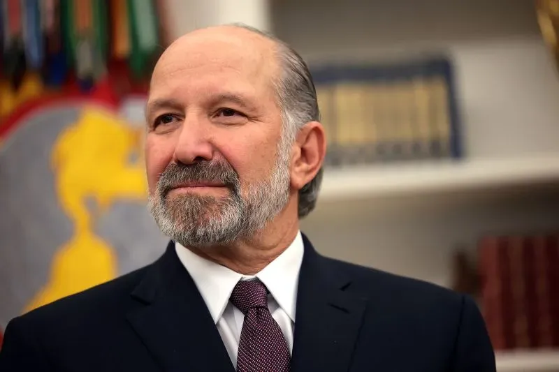 صورة لرجل ذو لحية وشعر رمادي، يرتدي بدلة رسمية، يبدو في وضعية جدية. تظهر خلفه أعلام ورموز حكومية، مما يعكس سياق العمل الحكومي.