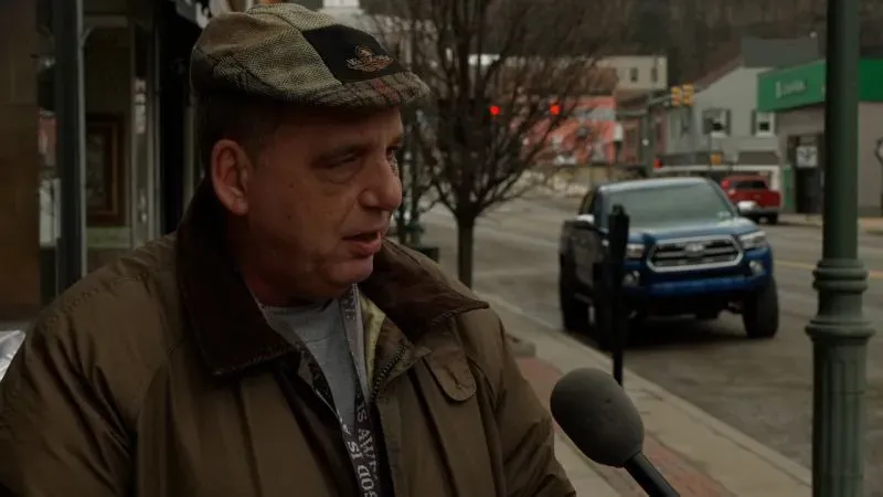 رجل يتحدث إلى ميكروفون في شوارع كيتانينغ، بنسلفانيا، مع خلفية لمتاجر محلية، يعبر عن آماله في تحسين الاقتصاد.