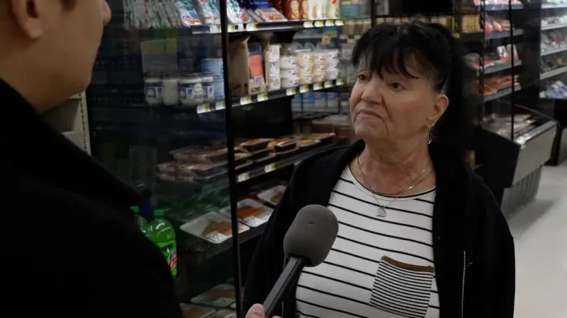 امرأة مسنّة تتحدث مع مراسل في متجر بقالة، محاطة بأرفف مليئة بالمنتجات الغذائية، تعبر عن قلقها بشأن ارتفاع الأسعار.