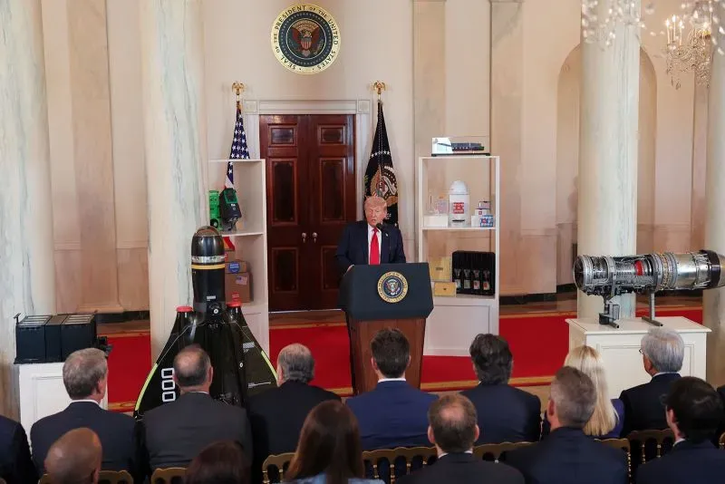 ترامب يتحدث في البيت الأبيض، مع عرض لمنتجات صناعية خلفه، في سياق مناقشة تأثير الحرب التجارية على الاقتصاد الأمريكي.