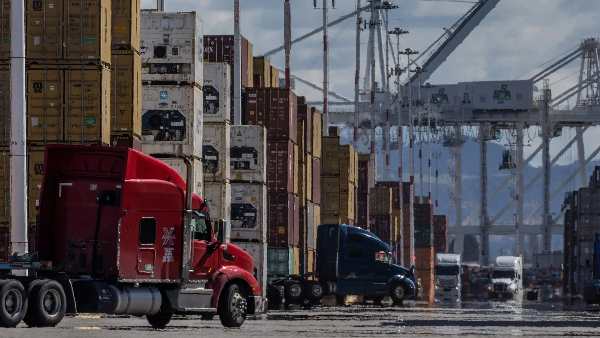مركبات شحن كبيرة متوقفة أمام حاويات بضائع مرتفعة في ميناء، تعكس حركة التجارة وتأثير التعريفات الجمركية.