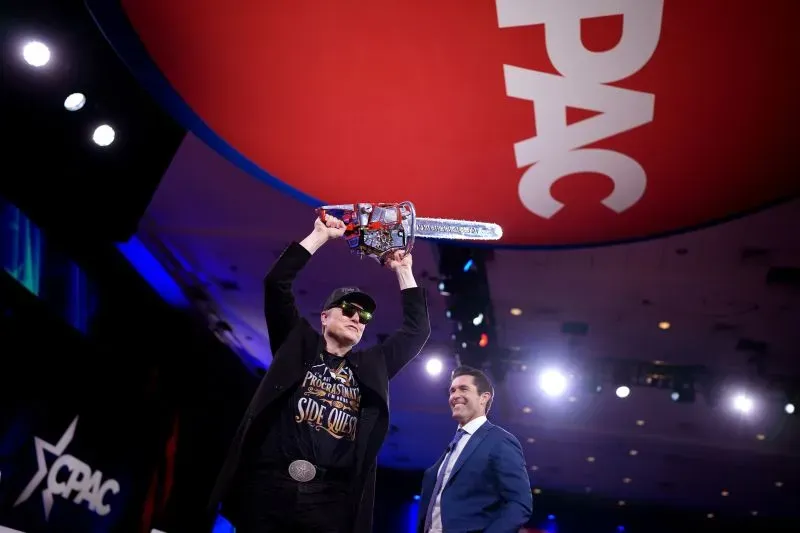 رجل يرتدي نظارات شمسية وقميصًا يحمل شعارًا، يرفع منشارًا كهربائيًا على خشبة المسرح خلال مؤتمر سياسي، بينما يقف رجل آخر مبتسمًا بجانبه.