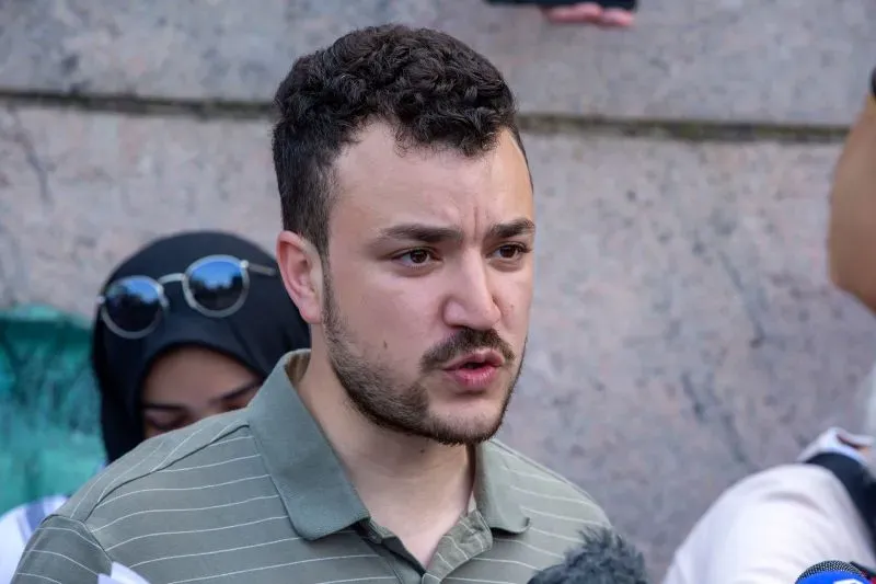 شخص يتحدث في تجمع عام، مع تواجد أشخاص آخرين في الخلفية، يعبر عن موقفه حول قضايا سياسية تتعلق بإدارة ترامب.