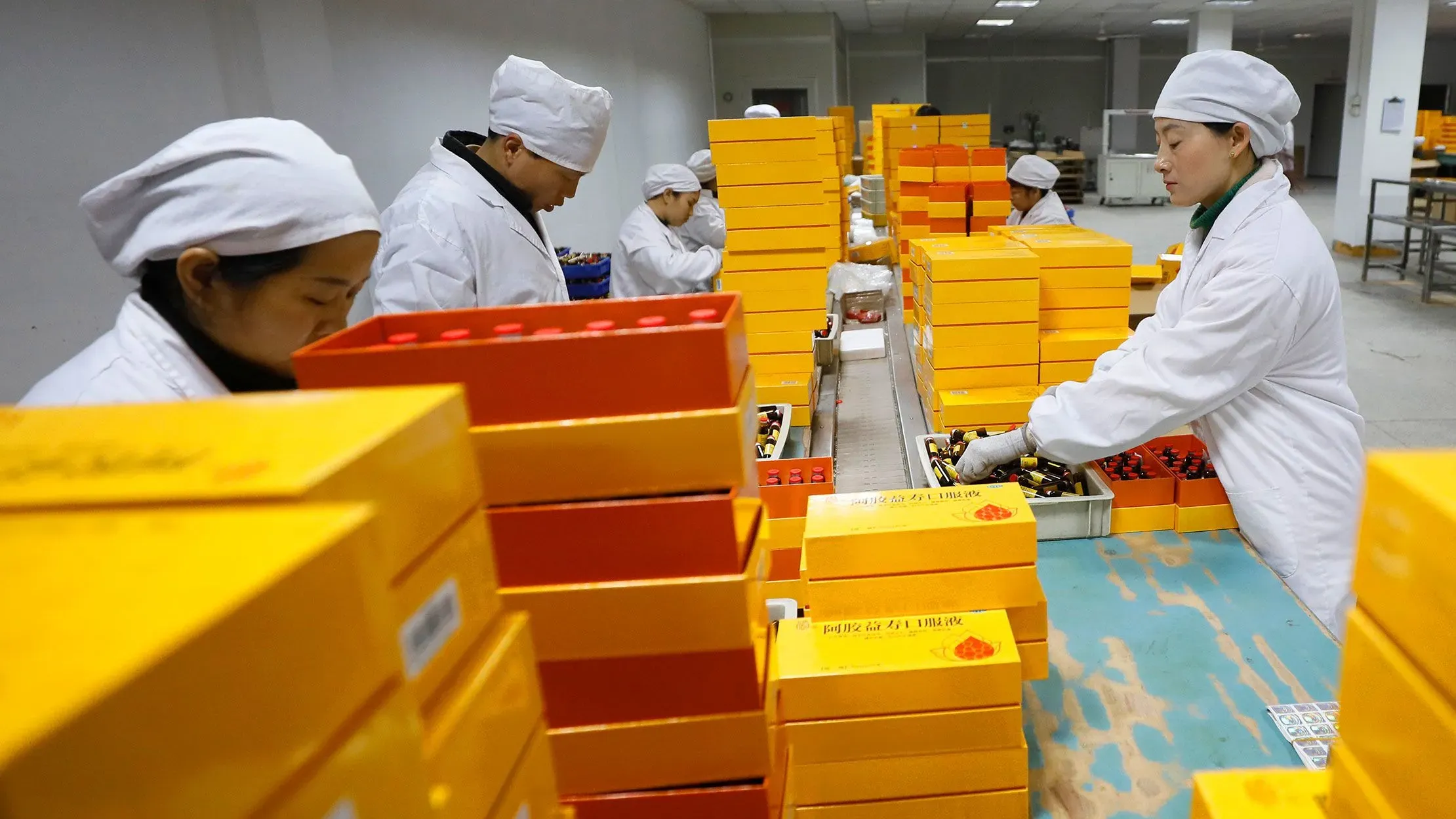 عمال في مصنع للأدوية يقومون بتعبئة صناديق الأموكسيسيلين، مع تكديس الصناديق الملونة في الخلفية، مما يعكس اعتماد أمريكا على الإنتاج الصيني.