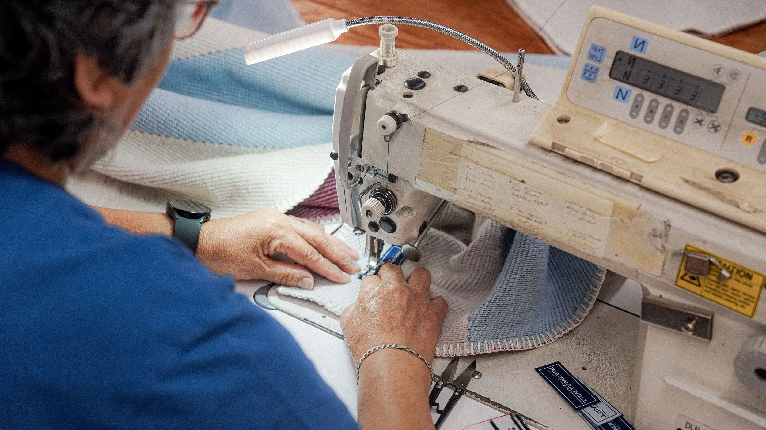عمال في مطحنة فاريبولت يقومون بخياطة بطانية باستخدام ماكينة خياطة حديثة، مع التركيز على الجودة والحرفية المحلية.