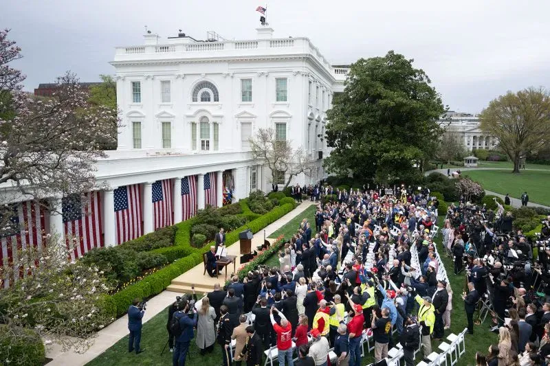 مؤتمر صحفي في حديقة البيت الأبيض بحضور حشد كبير، مع العلم الأمريكي يزين المبنى، يتحدث فيه الرئيس ترامب عن الرسوم الجمركية الجديدة.