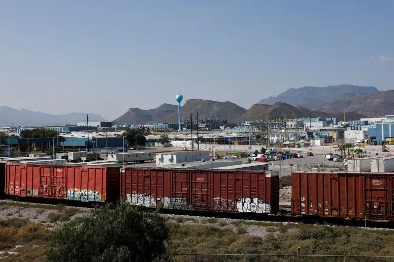 قطارات شحن تحمل بضائع، مع مصانع ومناطق جبلية في الخلفية، تشير إلى التبادلات التجارية بين الولايات المتحدة وكندا والمكسيك.