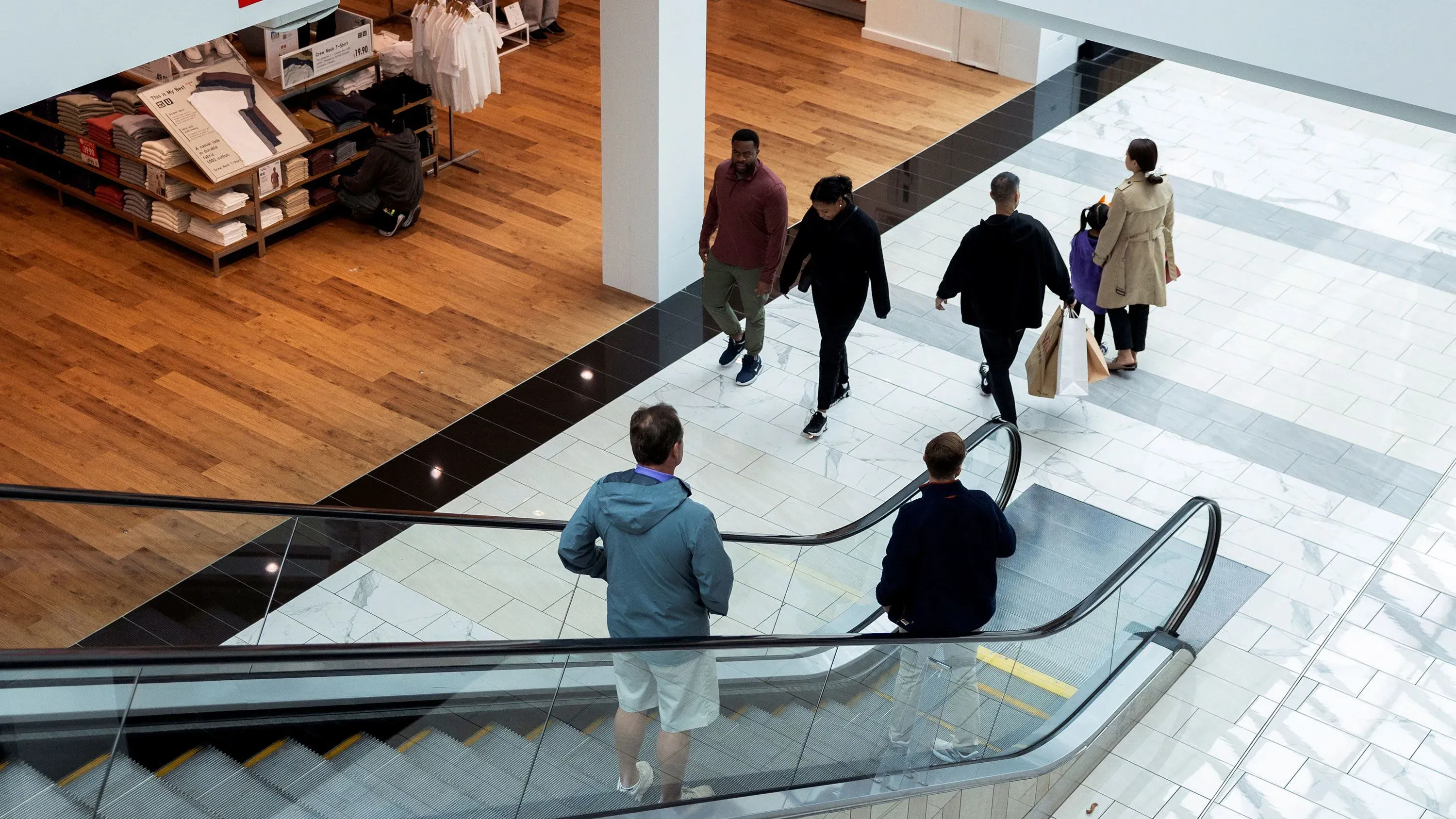 مجموعة من الأشخاص يتسوقون في مركز تجاري حديث، حيث يظهرون وهم يتنقلون بين المتاجر على سلالم متحركة، مما يعكس تأثير التعريفات الجمركية على خيارات المستهلكين.