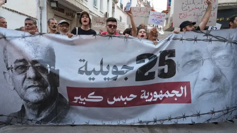 تظاهرة في تونس ضد الرئيس قيس سعيد، حيث يحمل المحتجون لافتة مكتوب عليها "الجمهورية سجن كبير" تنديدًا بالاستبداد السياسي.