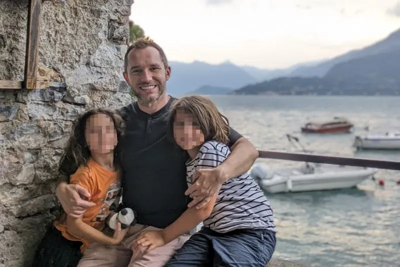 أب مع طفليه يجلسان بجانبه على ضفاف بحيرة، مع خلفية جبال ومراكب، يعكس اللحظات العائلية السعيدة في سياق مأساوي.