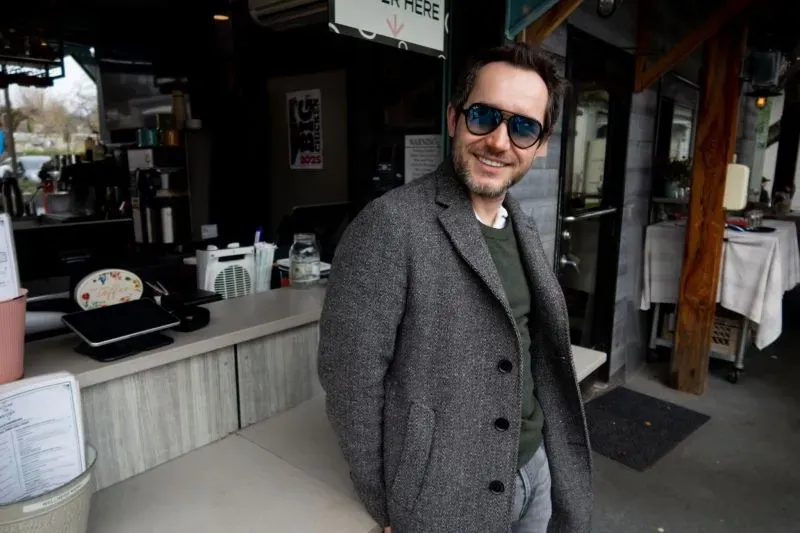 رجل مبتسم يرتدي نظارات شمسية ويقف أمام مقهى، يعكس أجواء غير رسمية، في سياق قصة تتعلق بقتل أستاذ تسويق في أثينا.