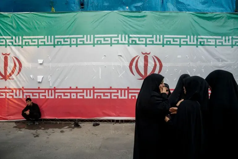 نساء يرتدين الحجاب الأسود يتحدثن معًا أمام علم إيران الكبير، في سياق التوترات السياسية المرتبطة بأنشطة الاستخبارات الإيرانية.