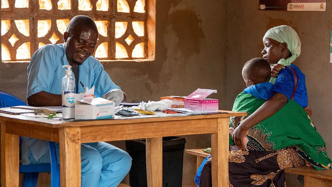 امرأة تحمل طفلًا في عيادة طبية، بينما يقوم طبيب بفحص الأدوية. تعكس الصورة تحديات مكافحة الملاريا في الكونغو.