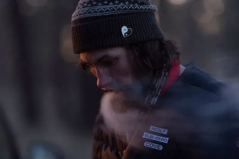 شخص يرتدي قبعة داكنة ويخرج بخارًا من فمه في غابة، يعكس أجواء القلق والتوتر بين المشردين بعد إغلاق المنطقة.