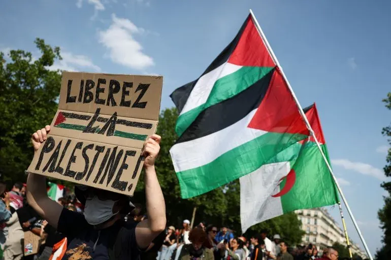 محتج يحمل لافتة تطالب بتحرير فلسطين، بينما ترفرف الأعلام الفلسطينية والجزائرية في الخلفية، خلال تظاهرة تضامن.