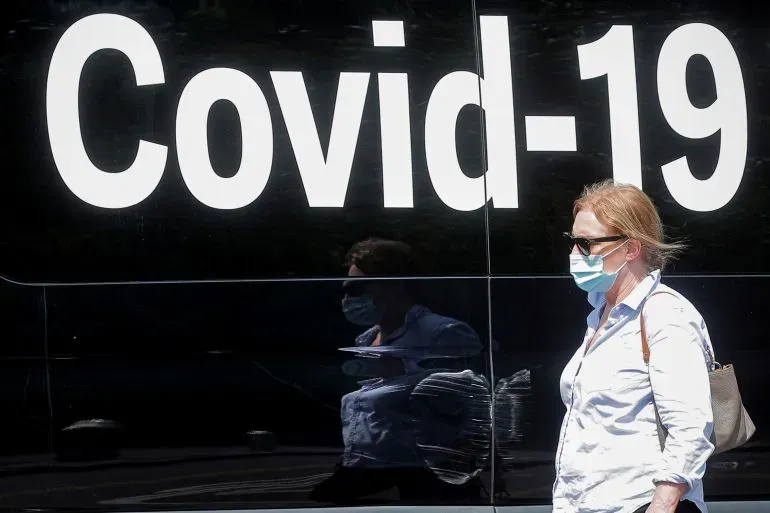 امرأة ترتدي كمامة تسير بجانب حافلة تحمل شعار "Covid-19"، في سياق مناقشات حول استجابة العالم للجائحة.