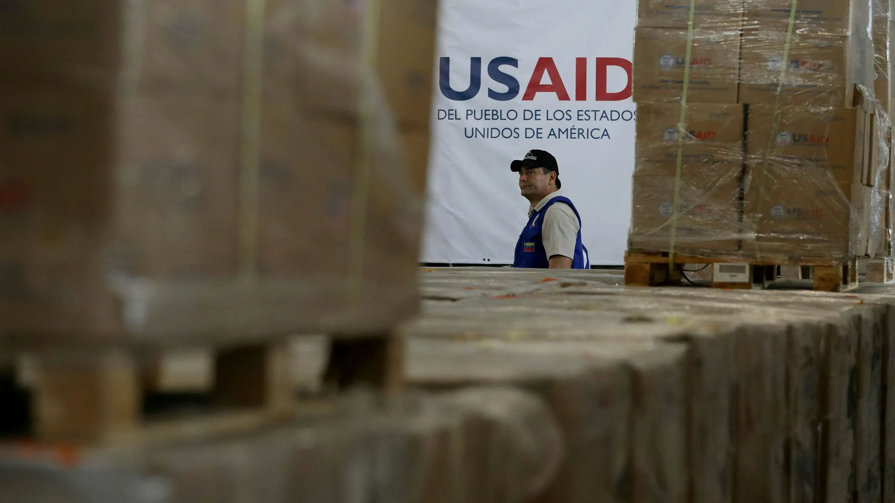 موظف في الوكالة الأمريكية للتنمية الدولية (USAID) يقف أمام لافتة الوكالة وسط صناديق مساعدات، في سياق مناقشات حول مستقبل الوكالة.