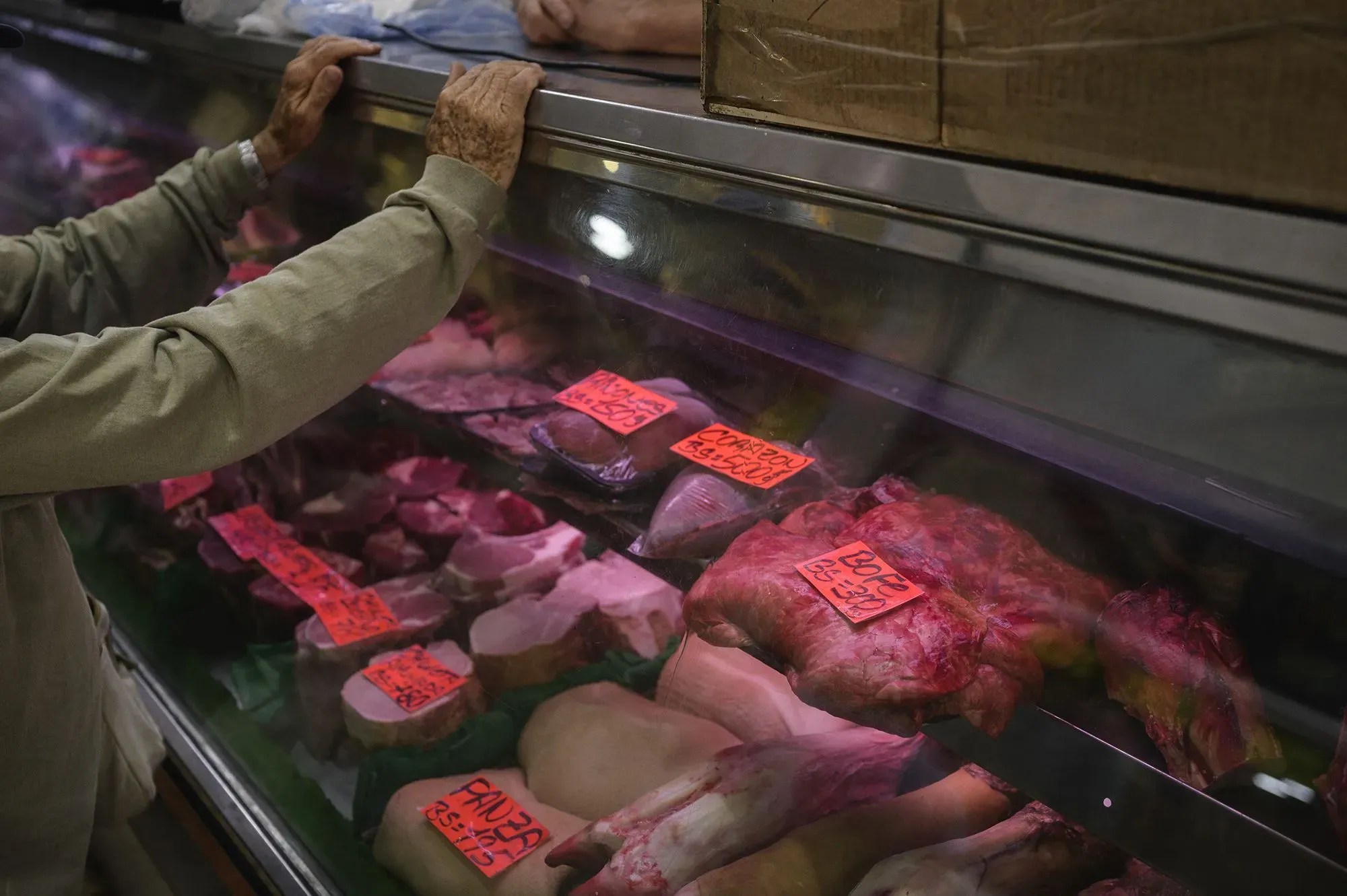 رجل مسن يتفحص اللحوم المعروضة في محل بقالة بفنزويلا، حيث تظهر الأسعار المرتفعة على الملصقات، مما يعكس أزمة الغذاء.