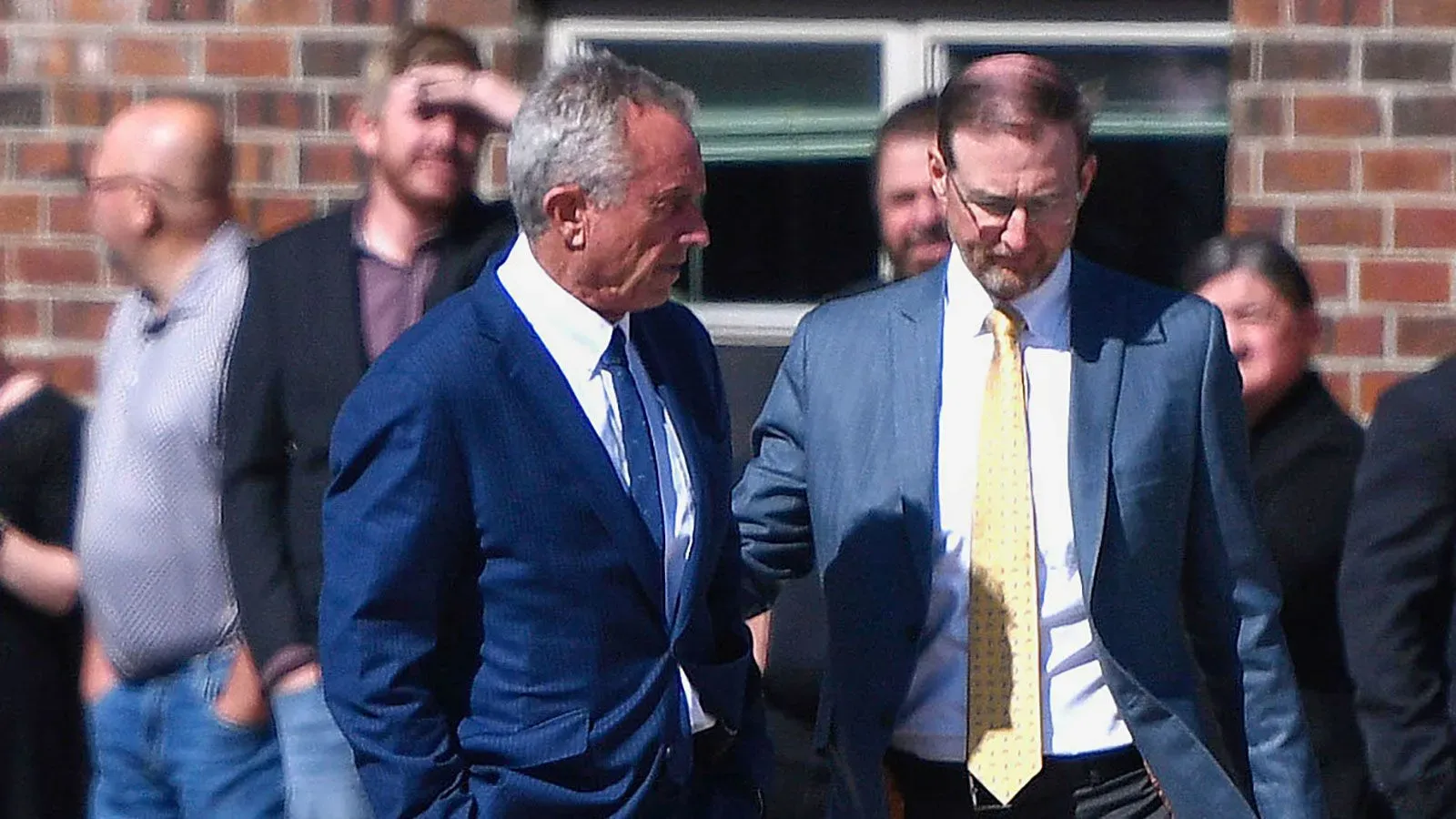 رجلان يمشيان معًا في الشارع، أحدهما يرتدي بدلة زرقاء ويبدو أنه يتحدث بجدية، بينما الآخر يرتدي بدلة رمادية.