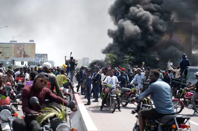 تظهر الصورة مشهدًا حيويًا للاحتجاجات في كينشاسا، حيث يتجمع المتظاهرون وسط دخان كثيف، مع وجود أفراد الشرطة وعناصر في الخلفية.