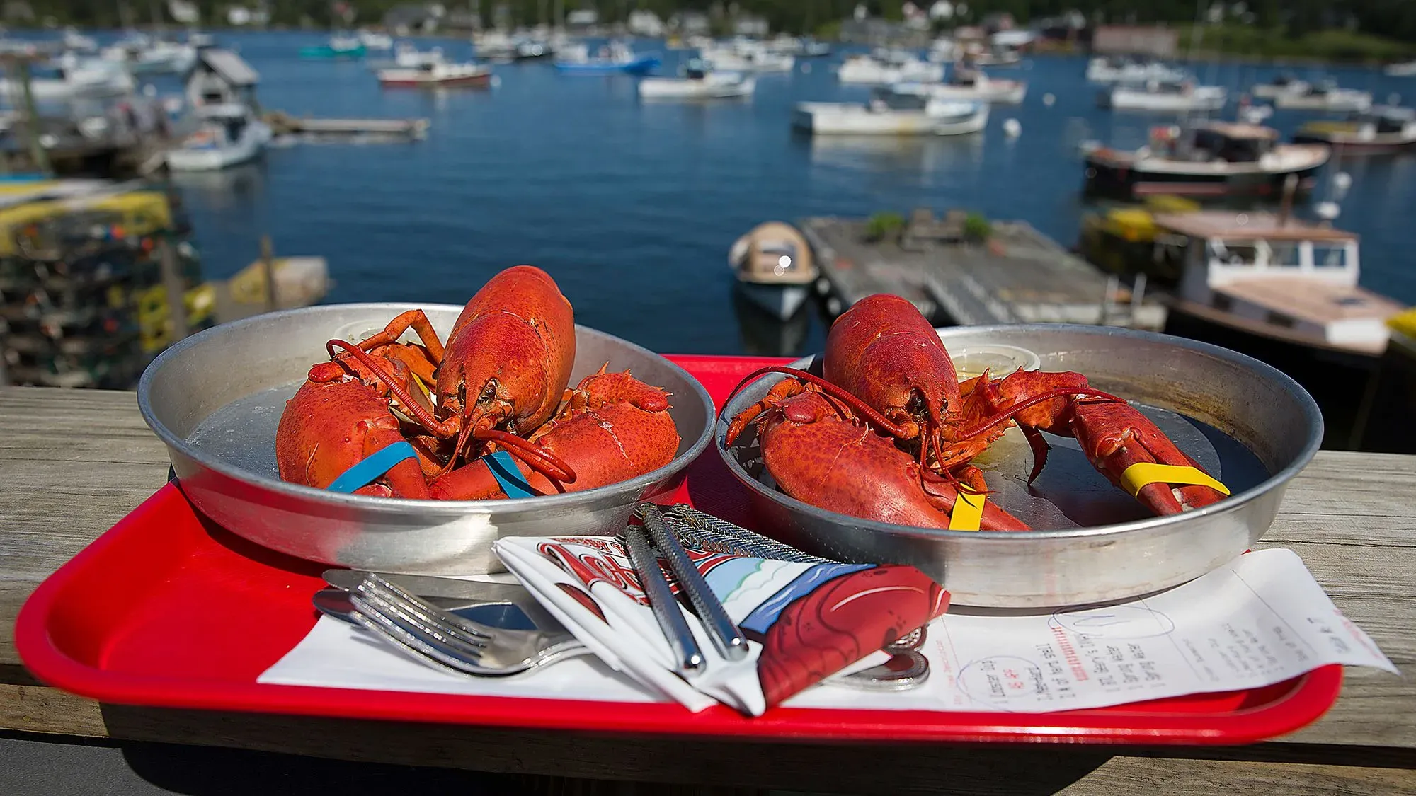 طبق يحتوي على كركند في مين، مع أدوات تناول الطعام، وخلفية بحرية تضم قوارب، مما يعكس الثقافة البحرية للولاية.