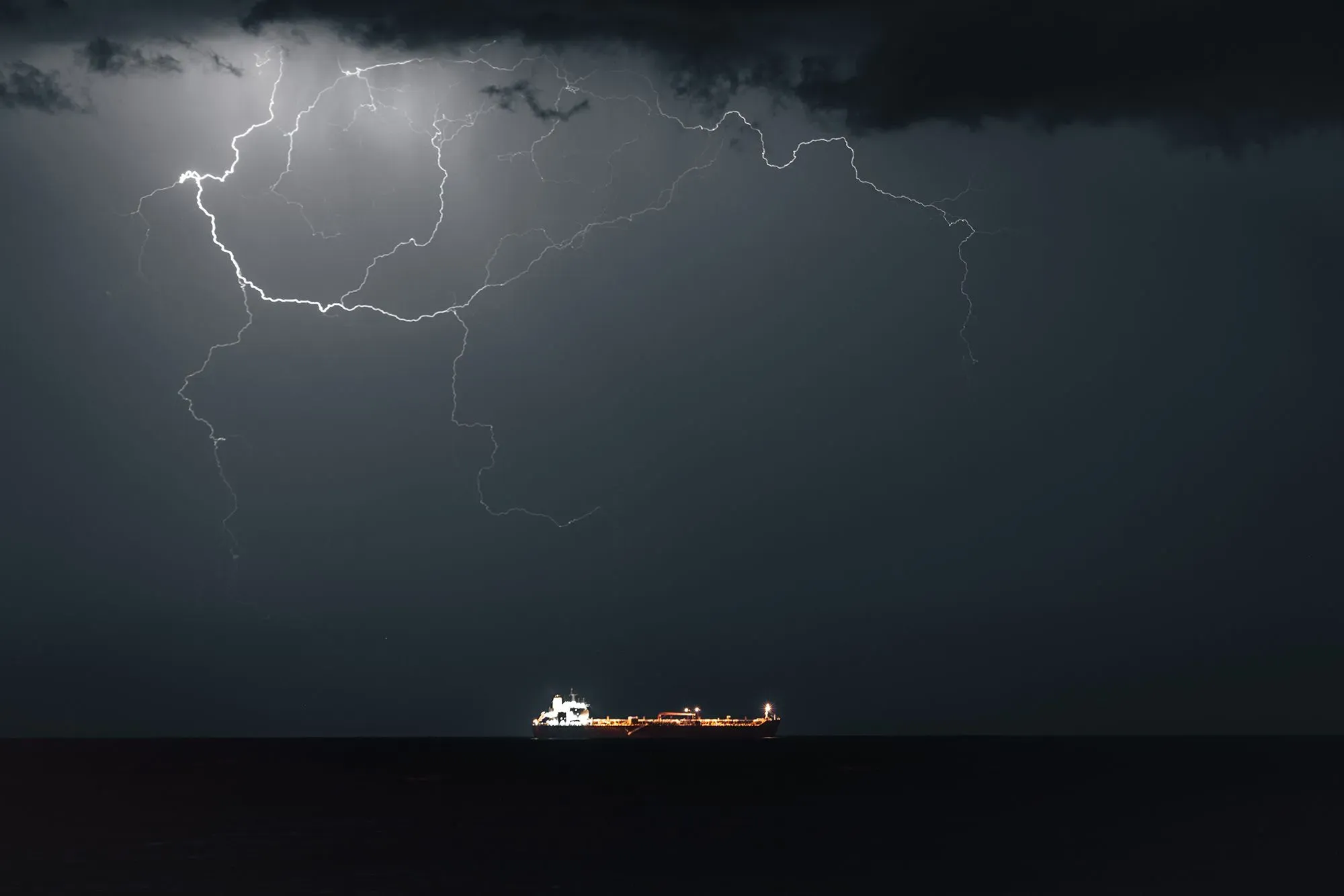 سفينة شحن تبحر في البحر تحت سماء ملبدة بالغيوم، مع وميض البرق في الخلفية، مما يعكس حالة عدم الاستقرار في مضيق هرمز.