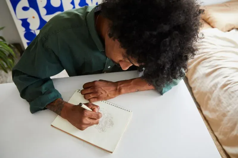 شخص يجلس على طاولة، يرسم في دفتر ملاحظات، مع التركيز على الإبداع كوسيلة للتغلب على القلق.