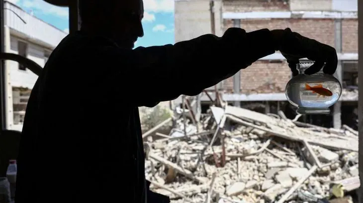 رجل يحمل وعاءً يحتوي على سمكة ذهبية، بينما تظهر خلفه أنقاض مبنى مدمر، مما يعكس تأثير الحرب على الحياة اليومية.
