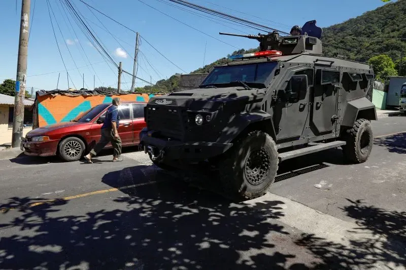 عربة مدرعة سوداء تجوب شوارع السلفادور، بينما يسير رجل بالقرب منها، مما يعكس الأجواء الأمنية المشددة في البلاد.