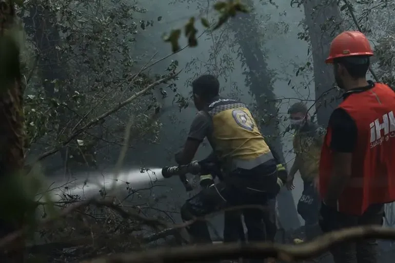 متطوعون في الدفاع المدني السوري يكافحون حرائق الغابات في مرتفعات اللاذقية، محاطون بالدخان والأشجار المحترقة.