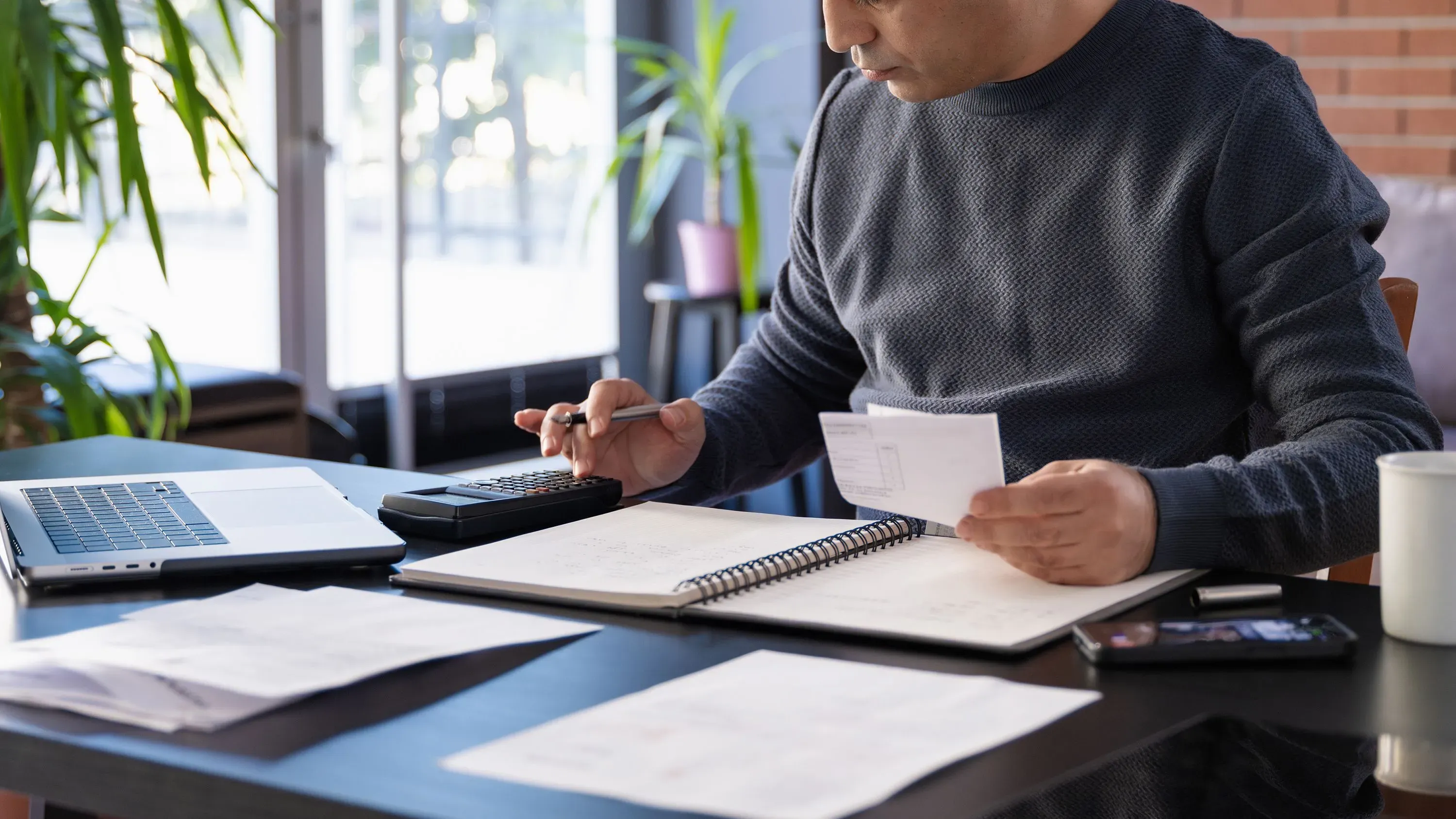 شخص يجلس على مكتب، يتفحص مستندات ضريبية ويستخدم آلة حاسبة، مع جهاز كمبيوتر محمول وملاحظات حول الضرائب لعام 2024.