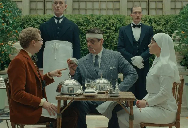 مشهد من فيلم "المخطط الفينيقي" لويس أندرسون، يظهر شخصيات تتناول الطعام في حديقة، مع اهتمام بالتفاصيل الأنيقة في الملابس.