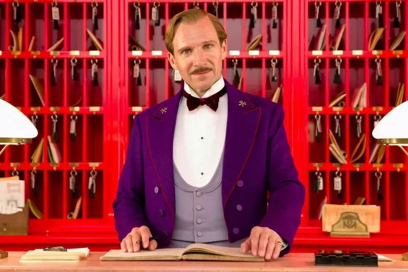مشهد من فيلم "The Grand Budapest Hotel" يظهر شخصية رئيسية ترتدي بدلة فاخرة بلون بنفسجي، أمام مكتب استقبال أنيق مع خلفية حمراء.