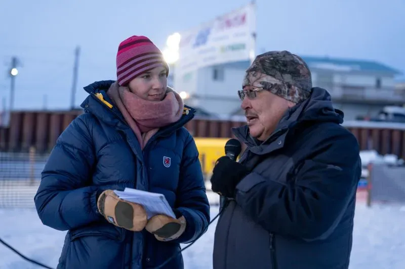 مراسل يتحدث مع رجل مسن في ألاسكا خلال بث إذاعي، مع خلفية لافتة إعلانات، مما يعكس أهمية الإعلام المحلي في المجتمعات القبلية.