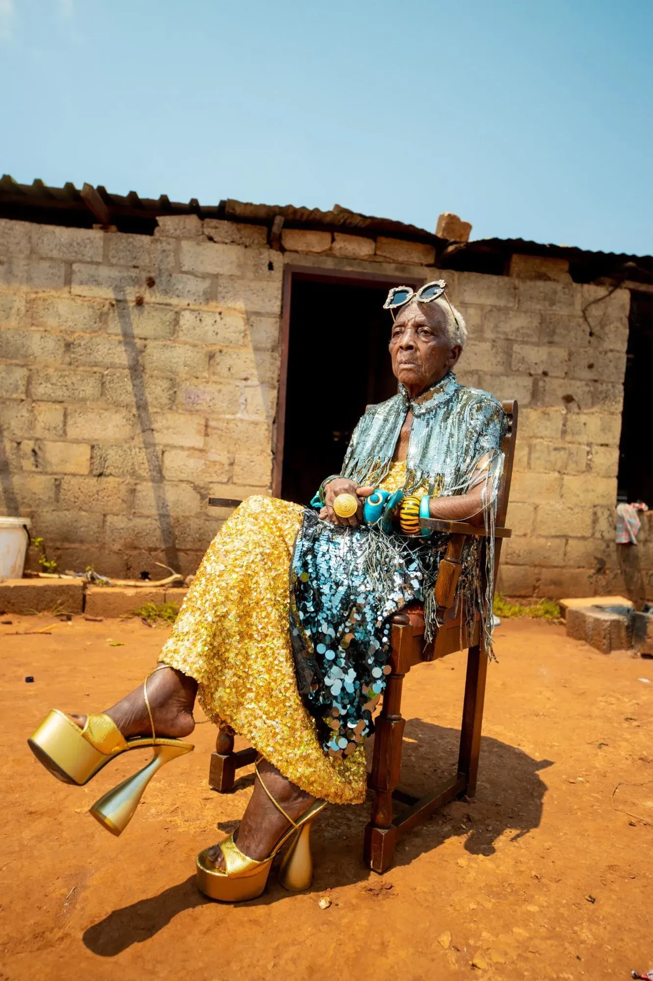 امرأة مسنّة ترتدي ملابس لامعة ومجوهرات، تجلس على كرسي أمام منزل بسيط، تعكس روح الأناقة والتحدي لفكرة الشيخوخة.