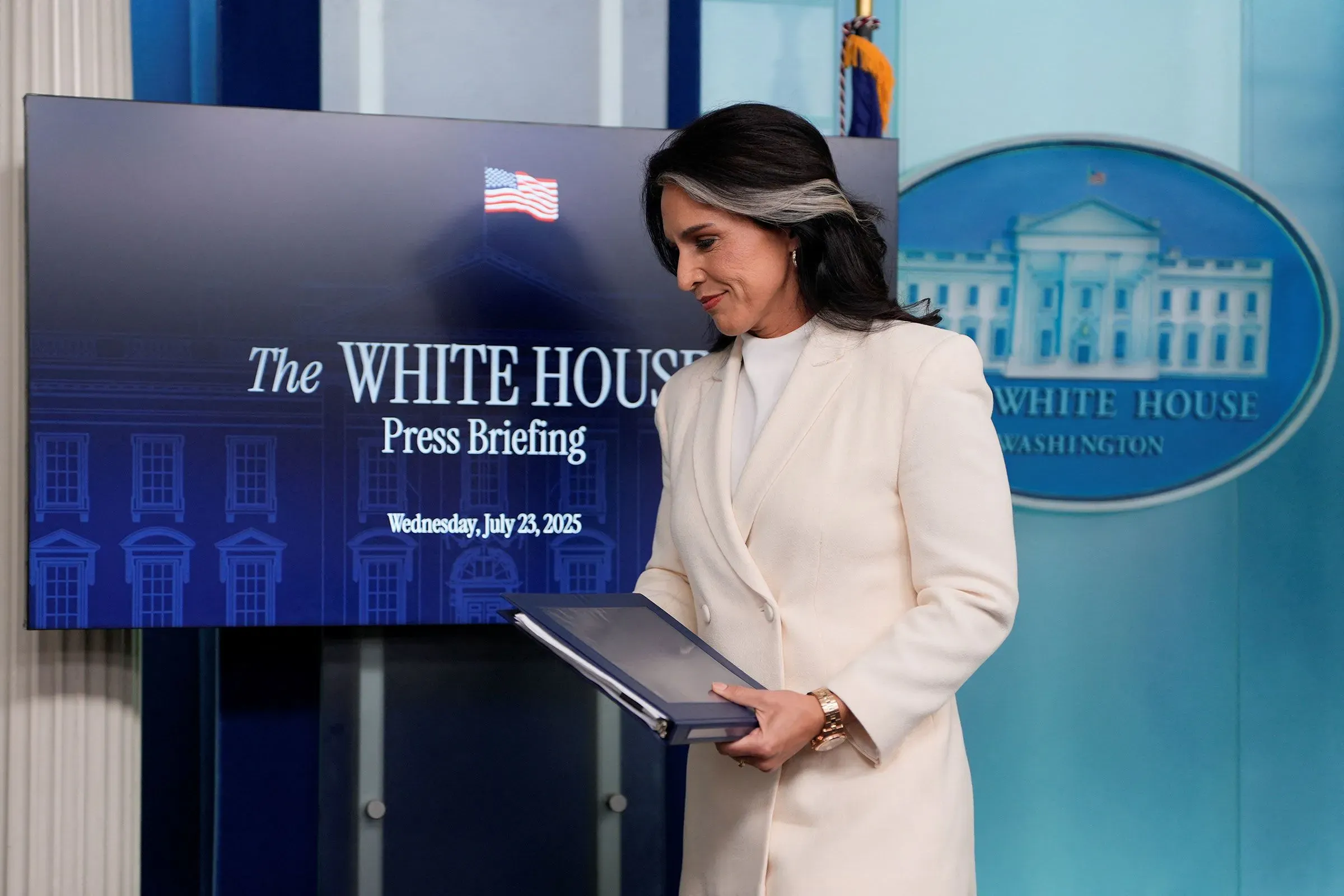تولسي غابارد أثناء مؤتمر صحفي في البيت الأبيض، تحمل جهاز كمبيوتر محمول، مع خلفية تتضمن شعار البيت الأبيض.