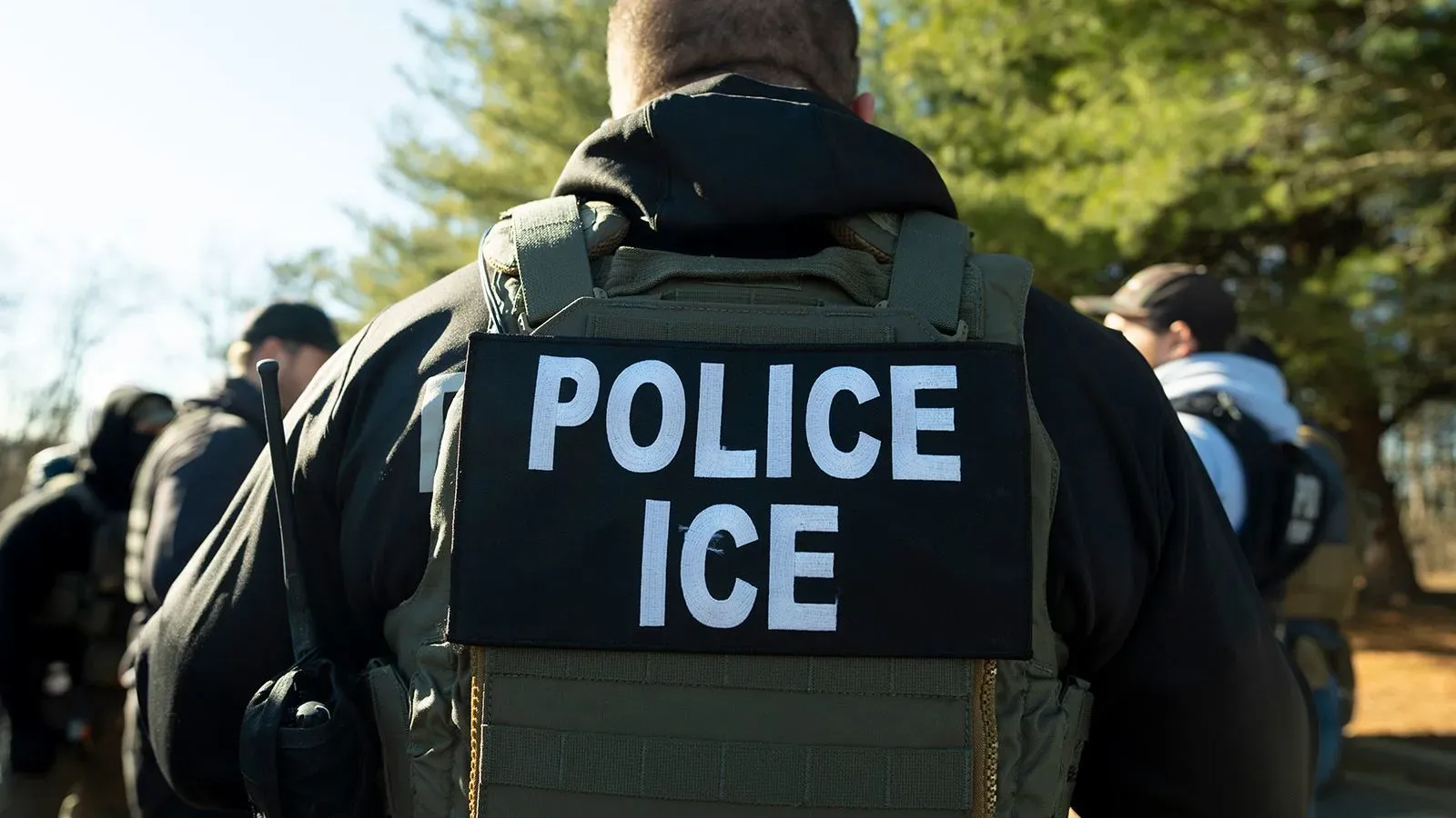 شرطي يرتدي زياً رسمياً يحمل علامة "شرطة ICE"، أثناء تنفيذ عمليات إنفاذ قوانين الهجرة في مكان خارجي.