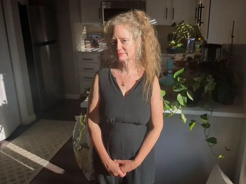 امرأة ترتدي فستاناً أسود وتقف في مطبخ مُضيء بأشعة الشمس، تعكس مشاعر الحزن والتأمل في التغيرات الناتجة عن حرائق الغابات.