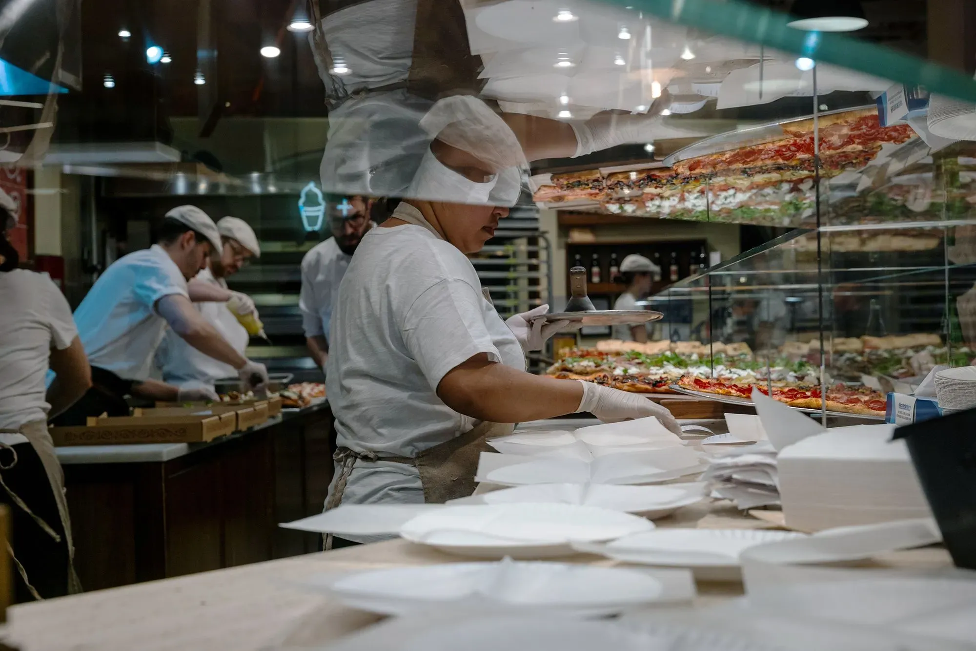 موظف في مطعم يقوم بتحضير الطعام، مع وجود زملاء في الخلفية، ويظهر تنوع الأطباق. الصورة تعكس نشاط قطاع الضيافة في سوق العمل.