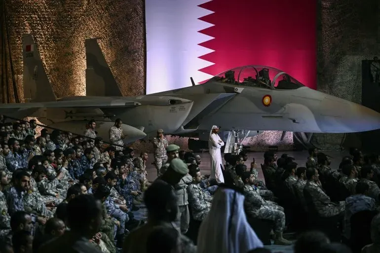 عرض عسكري في قطر يظهر طائرة مقاتلة من طراز F-15، مع علم قطر خلفها، وجنود يرتدون الزي العسكري يجلسون في الصفوف الأمامية.