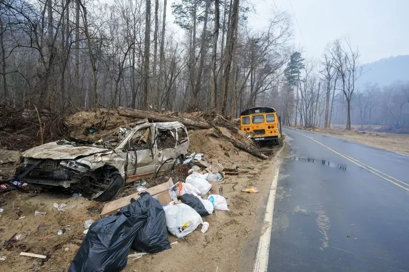 منطقة متضررة من الكوارث، تظهر سيارة مدمرة وأخرى قديمة على جانب الطريق، مع أكوام من النفايات والأشجار المتساقطة.