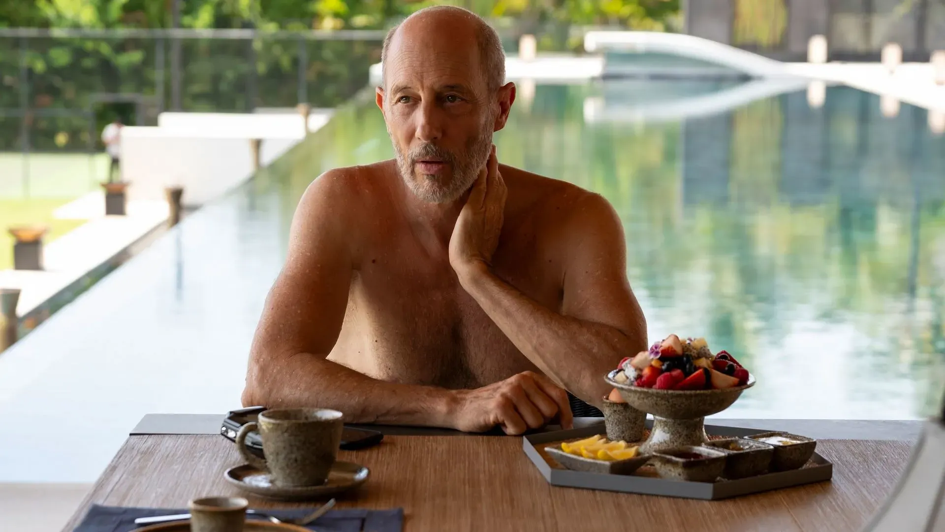 جريج هانت، شخصية مسلسل "اللوتس الأبيض"، يجلس بجانب حمام سباحة في منتجع بتايلاند، يتناول الفطور ويبدو عليه التفكير العميق.