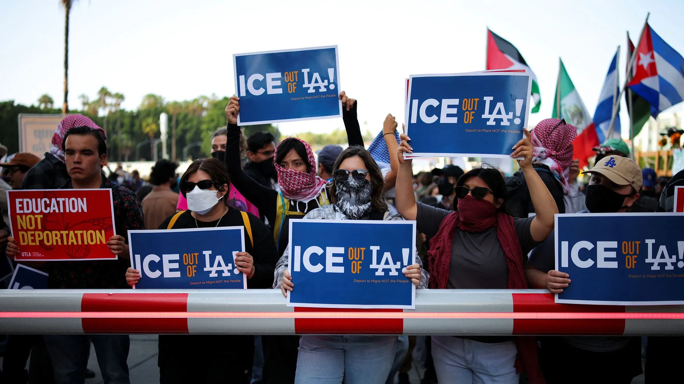 محتجون يحملون لافتات تطالب بإخراج إدارة الهجرة والجمارك من لوس أنجلوس، مع وجود أعلام تمثل ثقافات مختلفة في الخلفية.