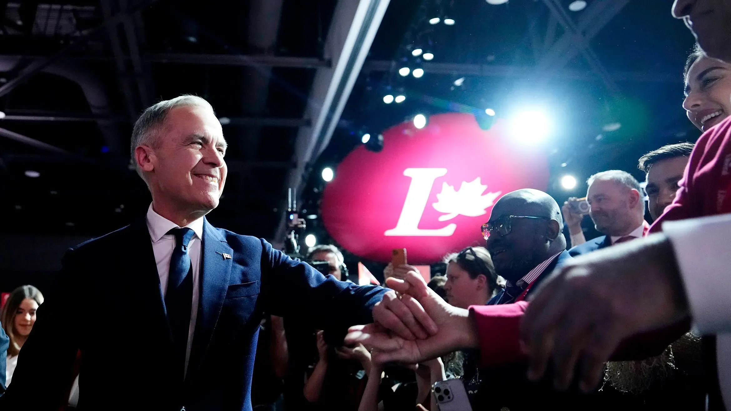 مارك كارني يبتسم ويصافح الحضور في حدث انتخابي للحزب الليبرالي الكندي، مع خلفية مضاءة بشعار الحزب.