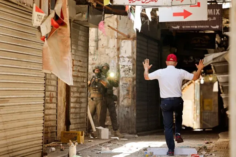 رجل يرفع يديه في شارع مغلق، بينما يراقبه جنود إسرائيليون. المشهد يعكس التوترات في الضفة الغربية تحت الحصار.