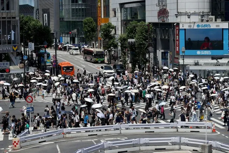 ازدحام مروري في شوارع طوكيو مع مجموعة من الأشخاص يحملون المظلات في يوم مشمس، مما يعكس التفاعل بين السكان والسياح.