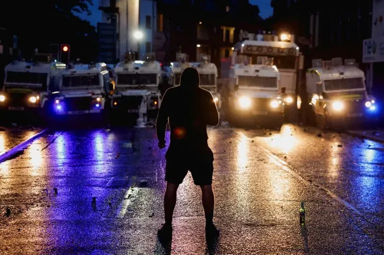 شخص يقف أمام صف من سيارات الشرطة المصفحة في أيرلندا الشمالية خلال اشتباكات احتجاجية، مع أضواء ملونة تعكس على الطريق.