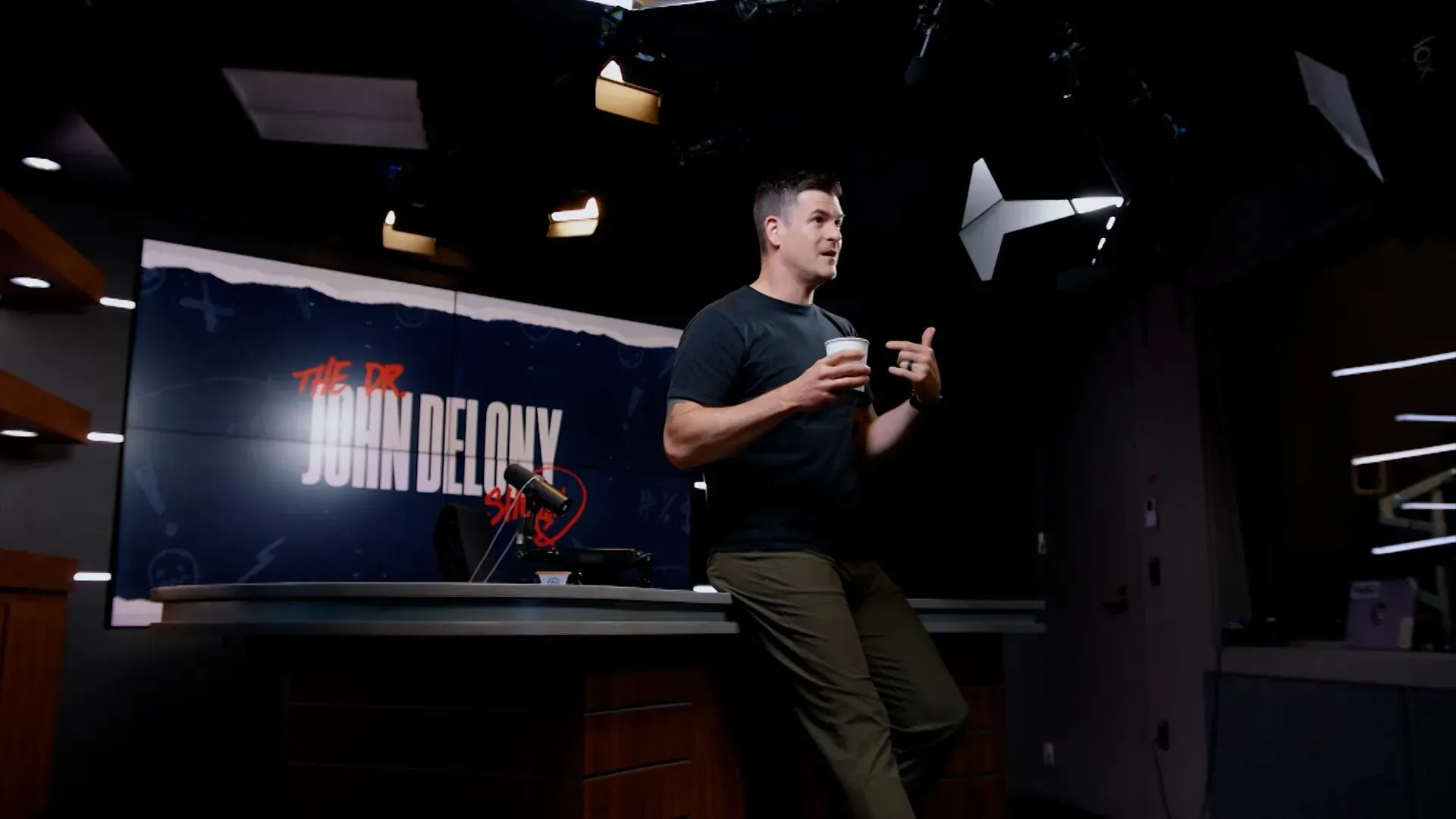 الدكتور جون ديلوني يتحدث إلى الكاميرا في استوديو البودكاست، ممسكًا بفنجان قهوة، مع خلفية تحمل اسم برنامجه.
