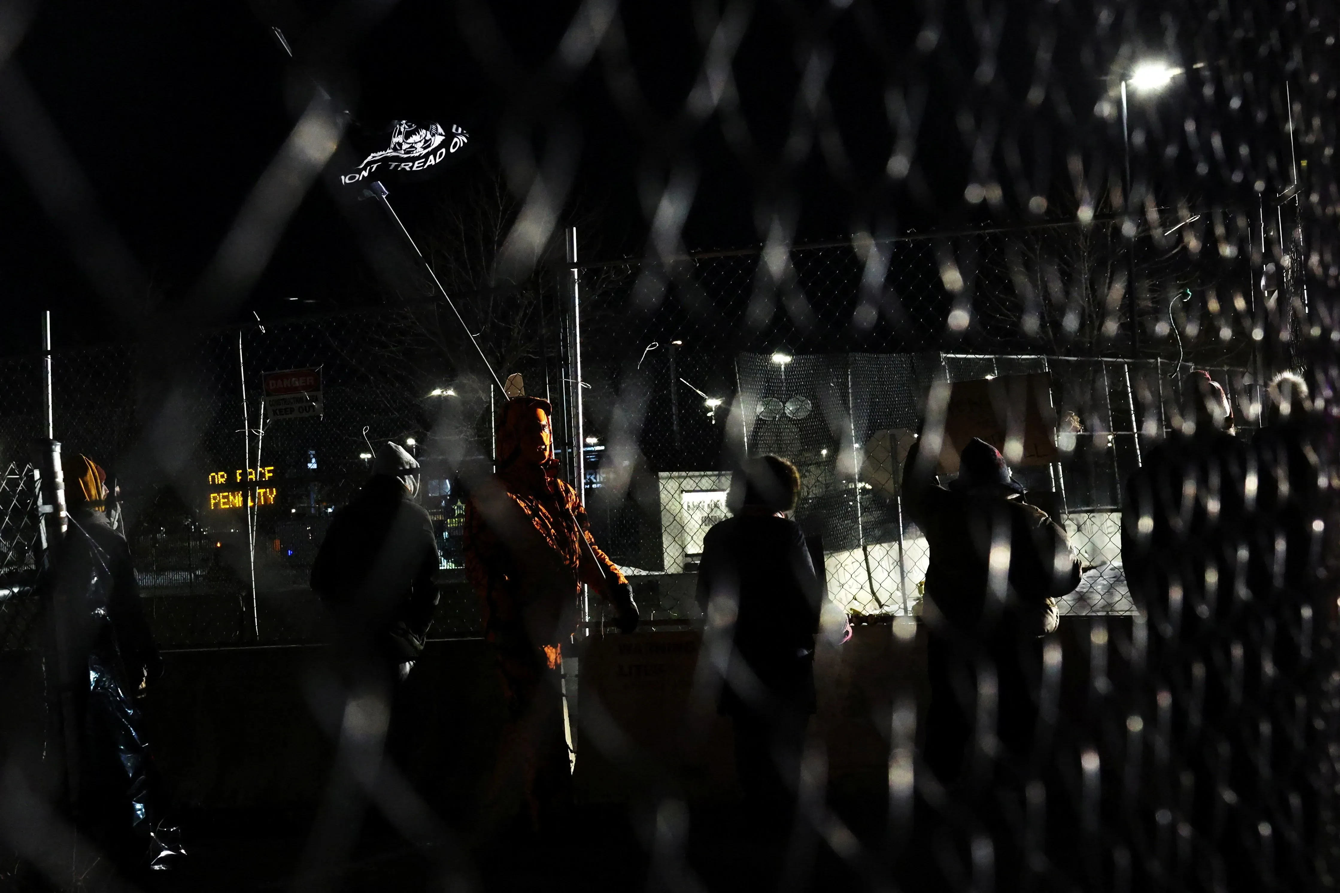 محتجون يقفون خلف سياج في مينيسوتا ليلاً، مع رفع علم يحمل شعار "لا تدوسوا علينا"، تعبيرًا عن الغضب من سياسات الترحيل الفيدرالية.